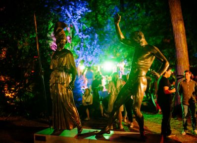Zwei Bronzeskulpturen auf einem Sockel bei Nacht in einem Garten, eine bekleidete Frau mit Helm und Lanze und ein nackter Mann, im Hintergrund buntes Licht und viele Menschen