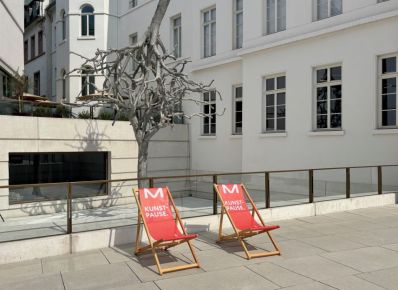 Zwei rote Liegestühle mit der Aufschrift Kunstpause auf einem Platz vor einem Gebäude