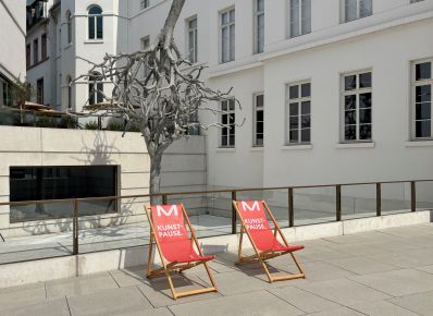 Zwei rote Liegestühle mit der Aufschrift Kunstpause auf einem Platz, dahinter ein weißes Gebäude