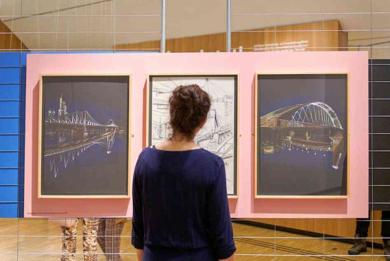 Ausstellungsansicht von zwei Fotos von Brücken und einer Zeichung in der Mitte vor der eine Frau steht und diese betrachtet