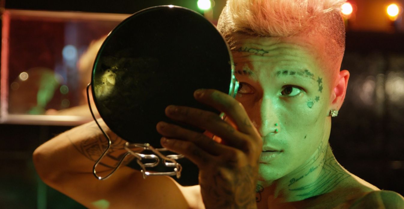 Enlarge image: Eine Person mit kurzen Haaren und Tatoos im Gesicht schaut sich in einem Handspiegel an