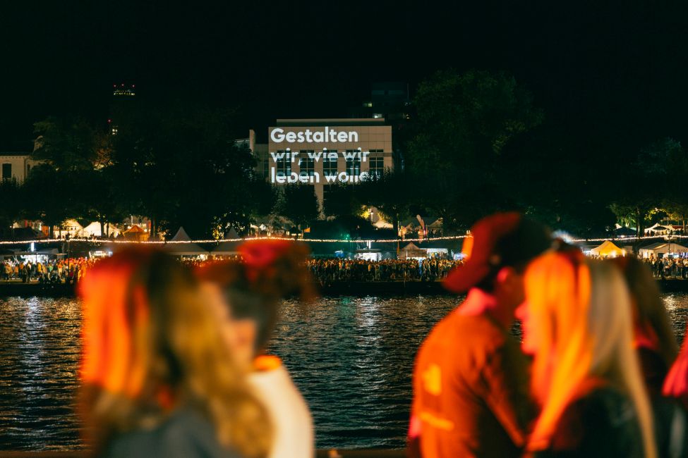 Enlarge image: Menschen spazieren bei Nacht am Flussufer, auf ein Haus ist 'Gestalten wir, wie wir leben wollen' projiziert