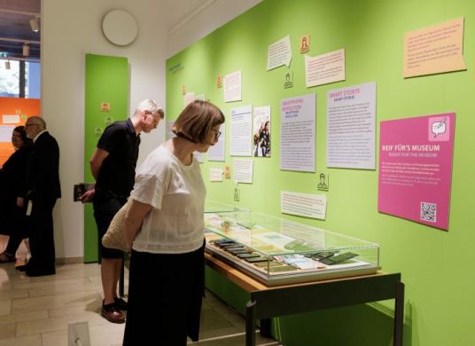 Menschen stehen in einem Ausstellungsraum mit Vitrinen und Texten an grünen Wänden und lesen